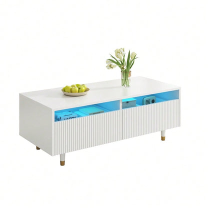 Dieser moderne LED-Couchtisch verfügt über eine gewellte Schubladenplatte, gold-weiße Beine, zwei Schubladen und einen farbwechselnden LED-Lichtstreifen. Ein passender TV-Schrank ist ebenfalls erhältl